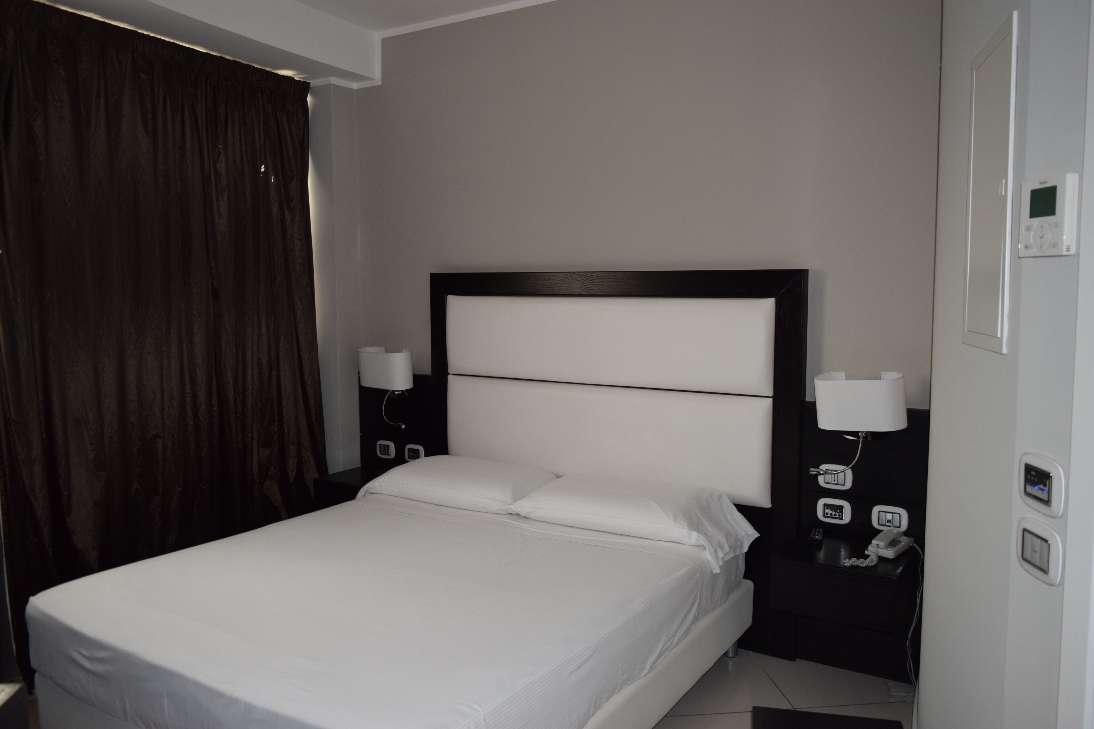 Habitación de hotel con una cama grande, una mesita de noche con lámpara y un termostato en la pared.