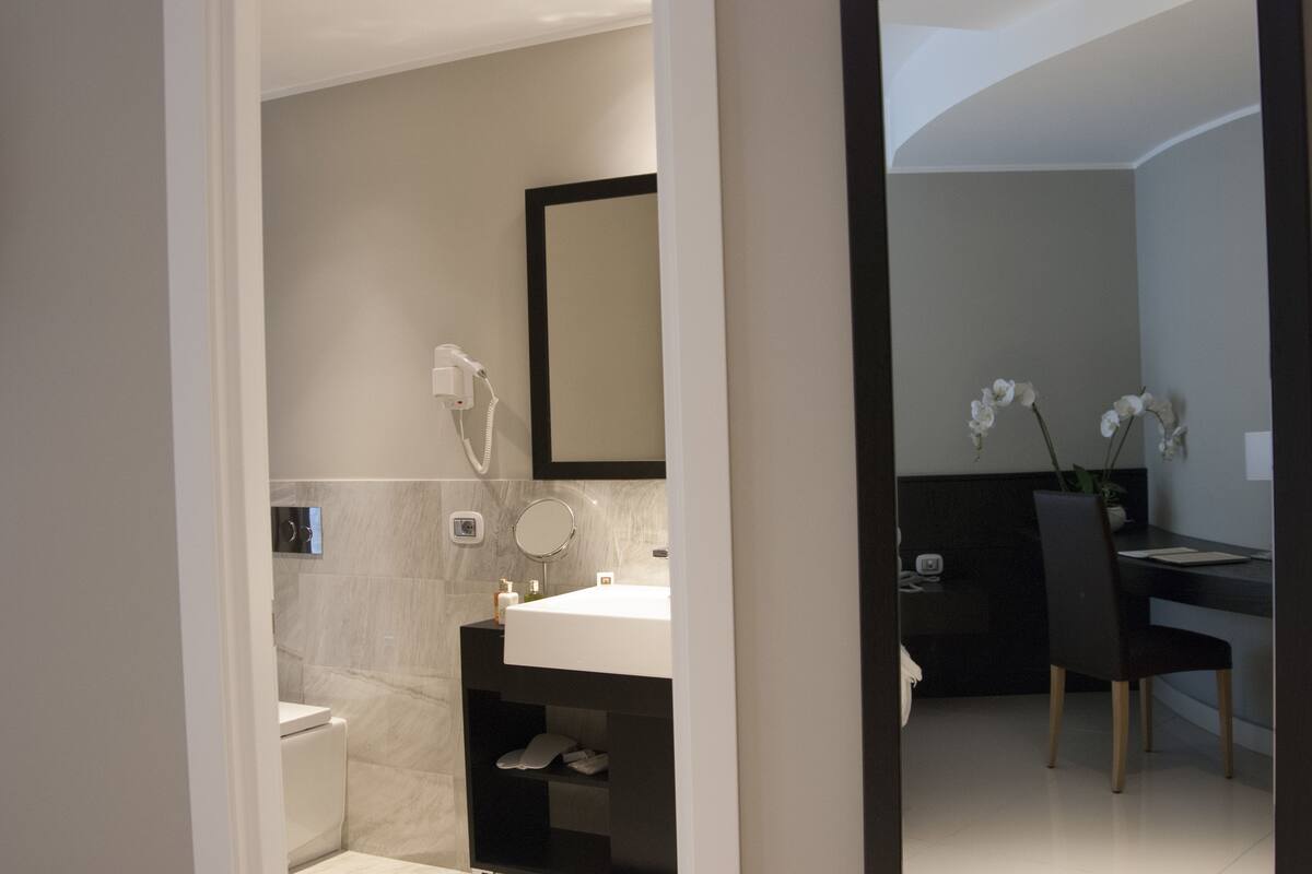 A modern bathroom with a toilet, sink, and shower. There is a mirror above the sink and a chair in the adjacent room.