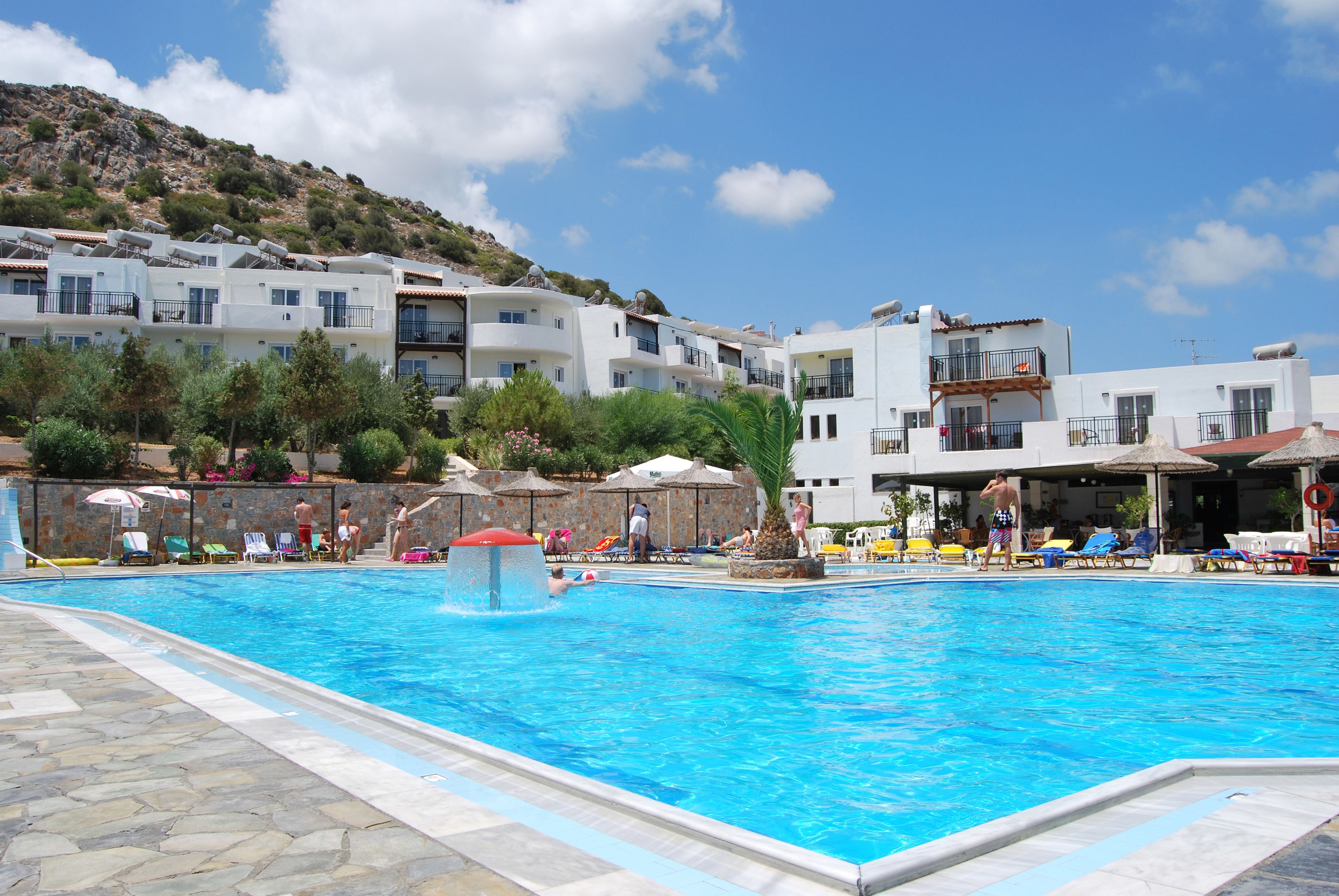 En swimmingpool med liggestole og parasoller, omgivet af hvide bygninger med balkoner og en bakke i baggrunden.