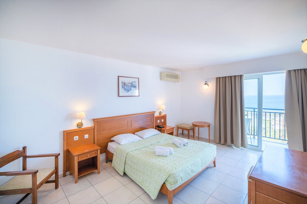A hotel room with a bed, wooden nightstands, a desk, a chair, and a balcony with curtains.