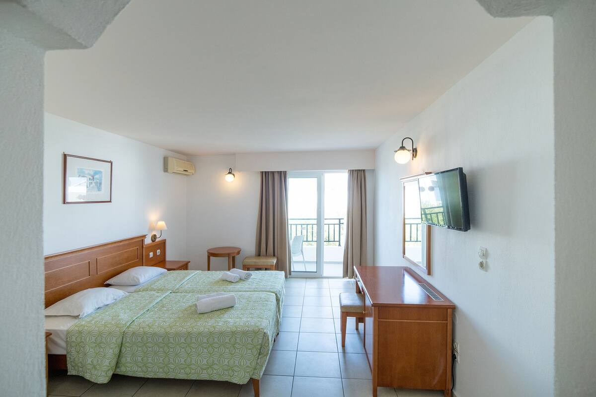 A hotel room with two beds, a desk, a TV, and a balcony with curtains.