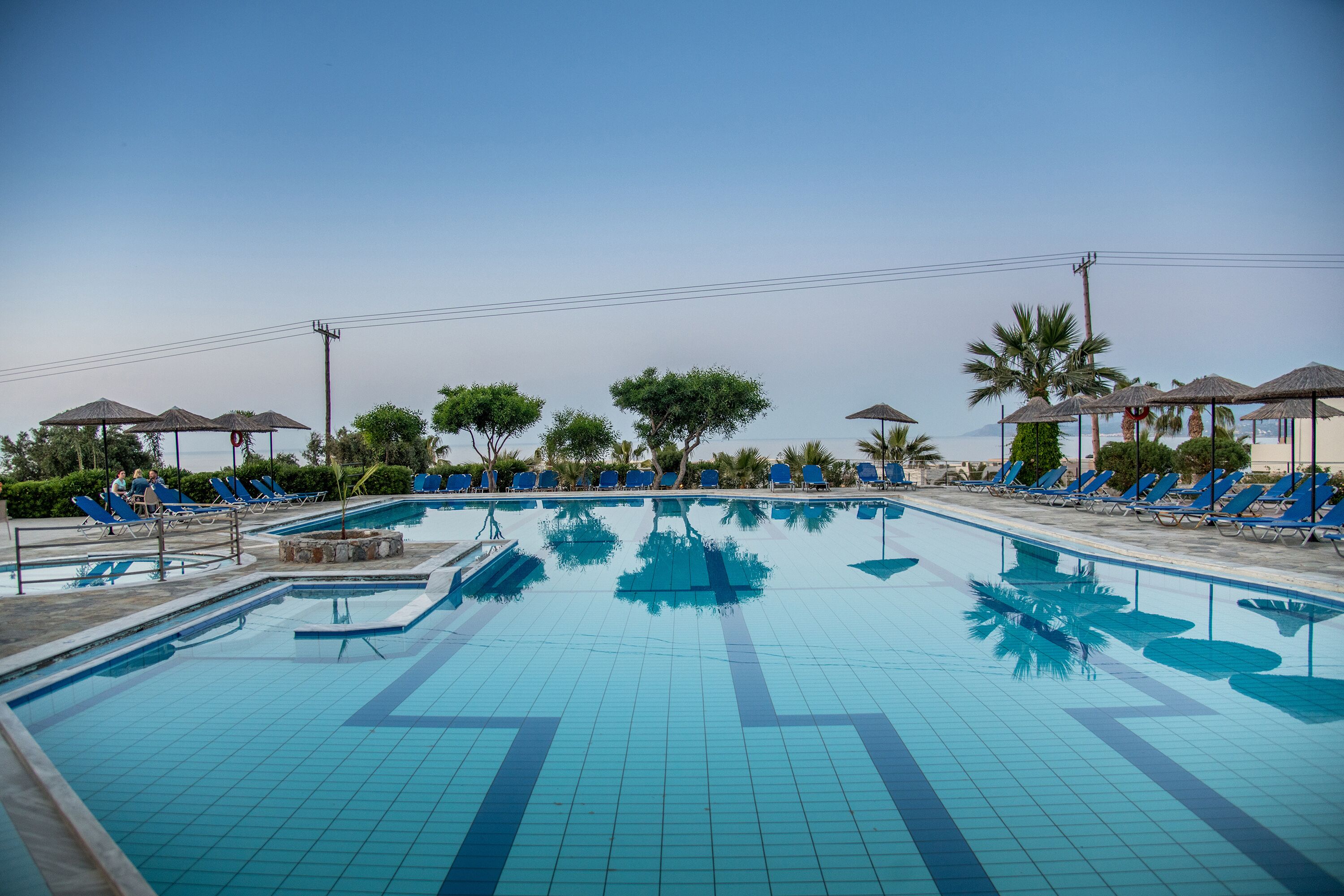 Una piscina con baldosas azules, rodeada de sillas de descanso y sombrillas, con árboles y un cielo despejado al fondo.