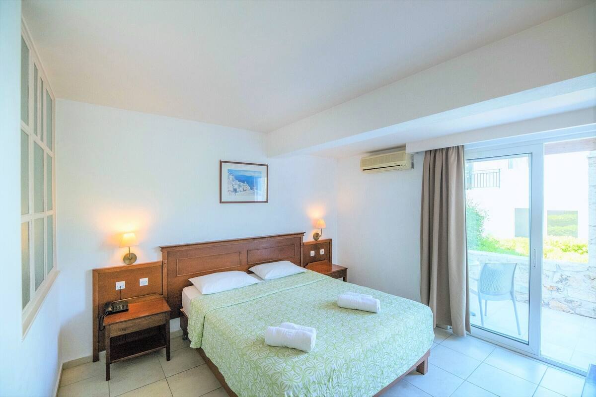 A hotel room with a bed, two bedside tables, a nightstand, a picture on the wall, and a sliding glass door to the outside.