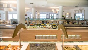 A buffet area with various food options, a seating area with tables and chairs, and a modern interior design with large windows and decorative plants. - Semiramis Village (Hersonissos)