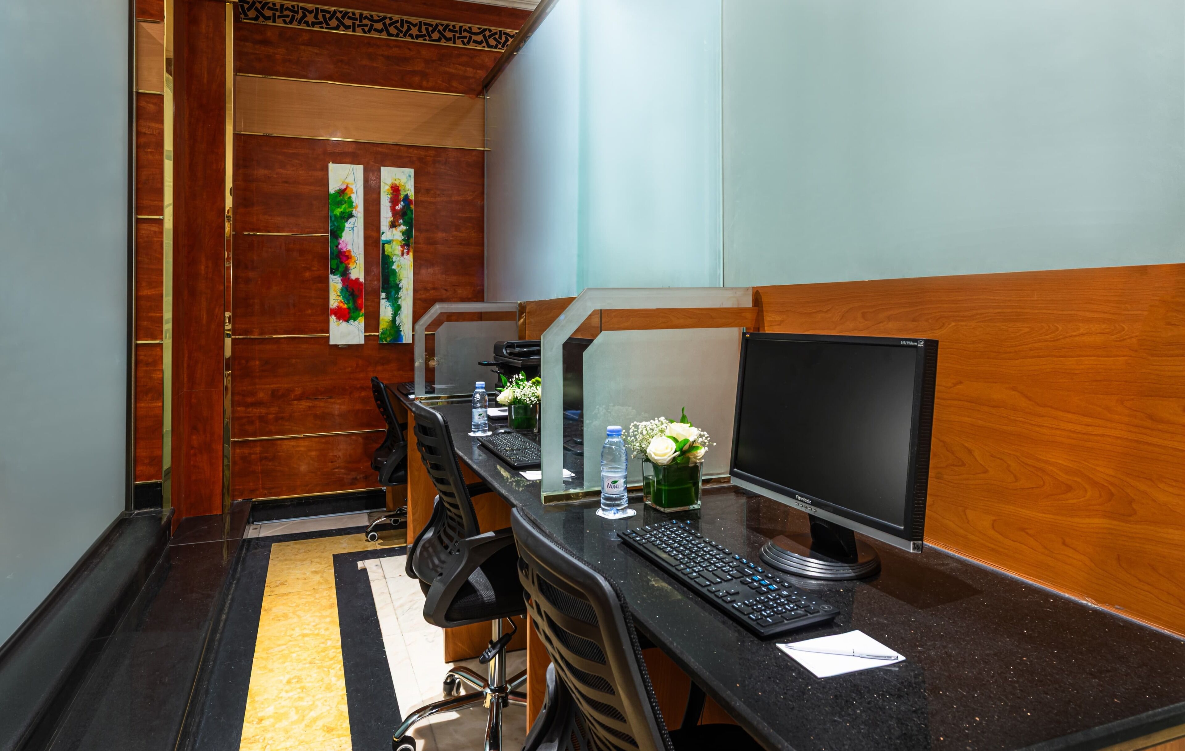 A hotel room with a long desk, multiple monitors, a keyboard, a water bottle, and a vase with flowers.