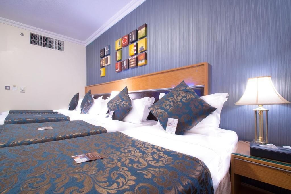 Une chambre d’hôtel avec un grand lit, plusieurs oreillers, une table de chevet avec une lampe et un mur orné de carreaux décoratifs.