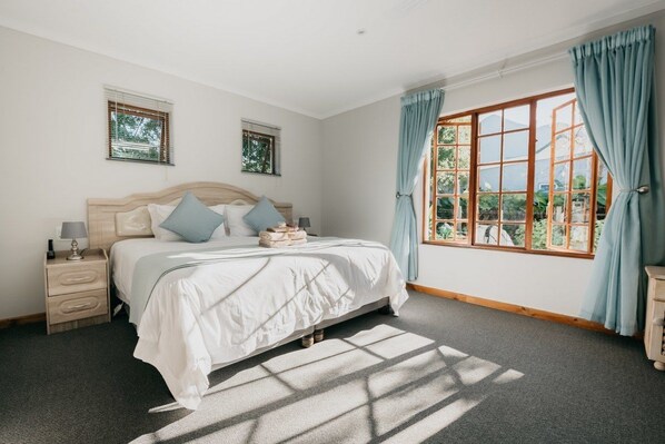 A bedroom with a large bed, two bedside tables, a nightstand, a window with curtains, and a view of greenery outside. - The Village Lodge (Storms River)