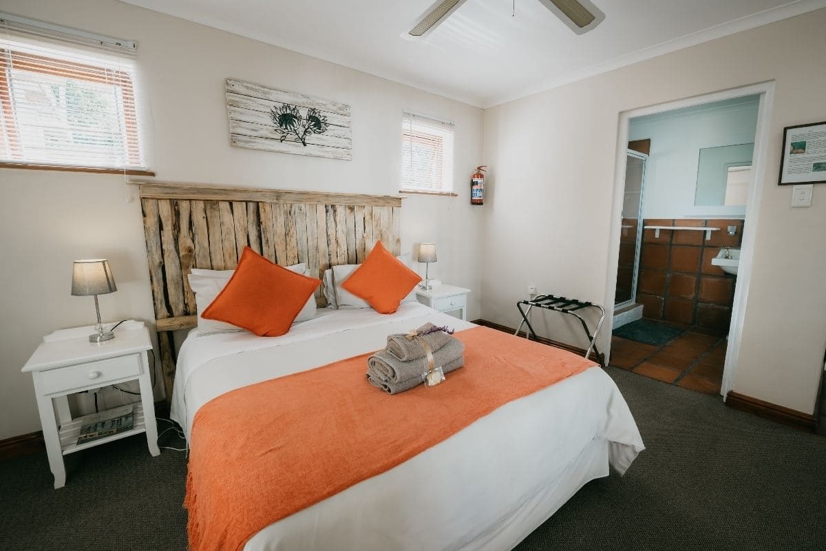 Habitación de hotel con cabecera de madera, dos mesitas de noche con lámparas, una manta color naranja y vistas al baño a través de una puerta abierta.