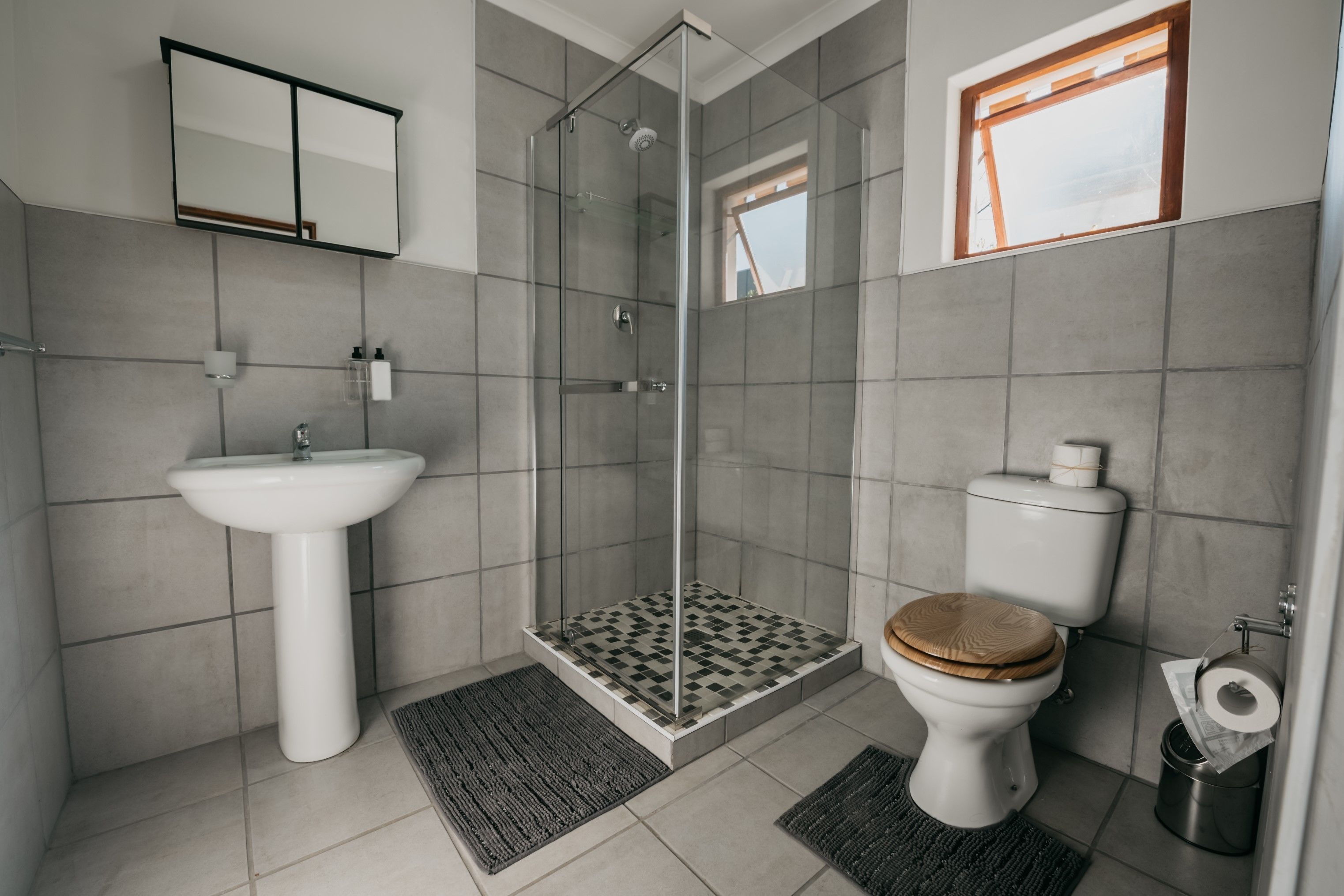 Un baño moderno con ducha envuelta en vidrio, lavamanos pedestal y inodoro con asiento de madera.