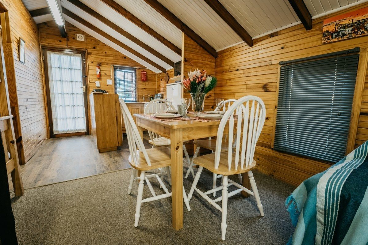 Interior de una cabaña de madera con una mesa de comedor dispuesta para cuatro, sillas blancas y una ventana con persianas.