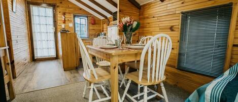 Interior de una cabaña de madera con una mesa de comedor dispuesta para cuatro, sillas blancas y una ventana con persianas.