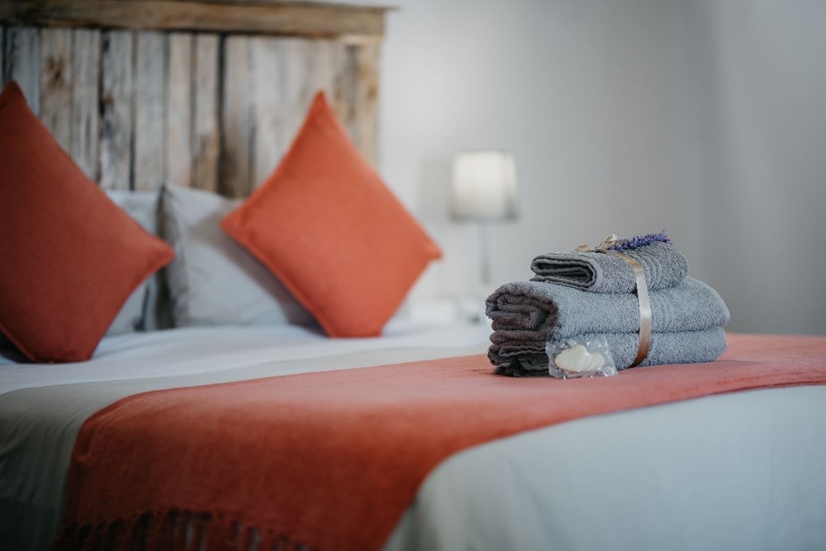 A neatly made bed with a red bedspread and pillows, a folded towel with a purple flower, and a rustic wooden headboard.