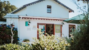 A white building with a red roof and a small window. There is a green awning and a sign on the door. - The Village Lodge (Storms River)