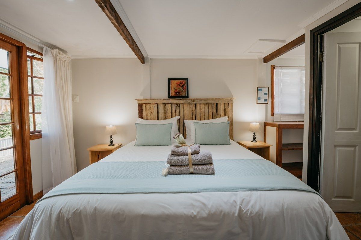 A neatly made bed with a light blue bedspread, two matching pillows, and a stack of folded towels. There is a wooden headboard, a small bedside table with a lamp, and a framed picture on the wall.