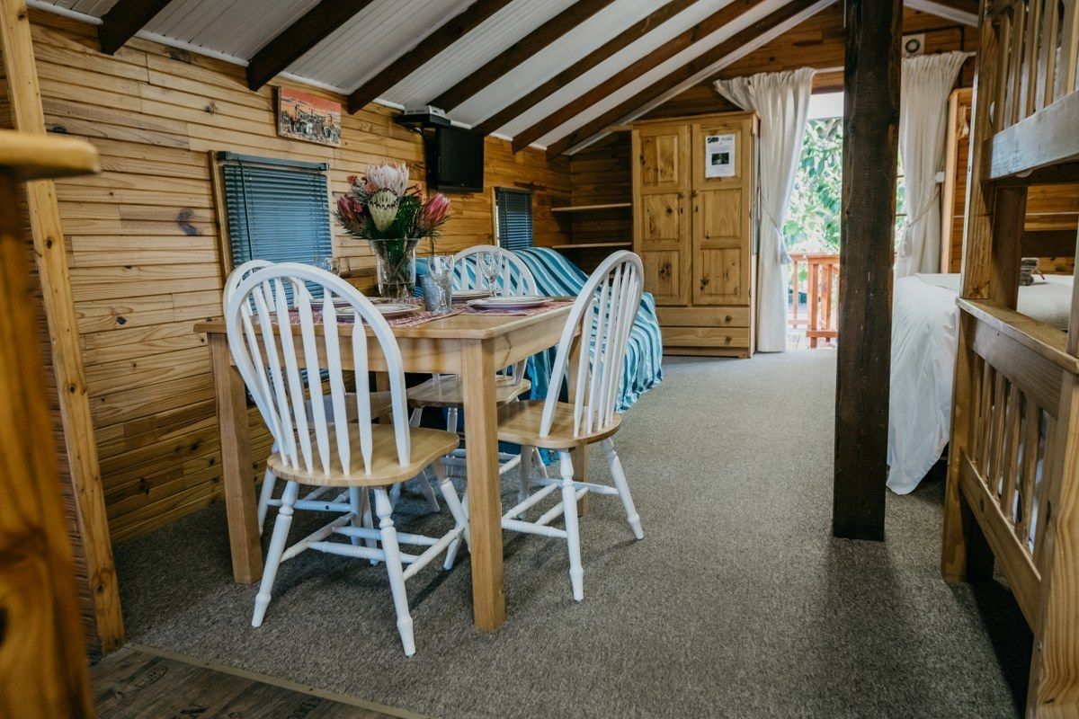 A wooden cabin interior with a dining table, white chairs, and a window with curtains.