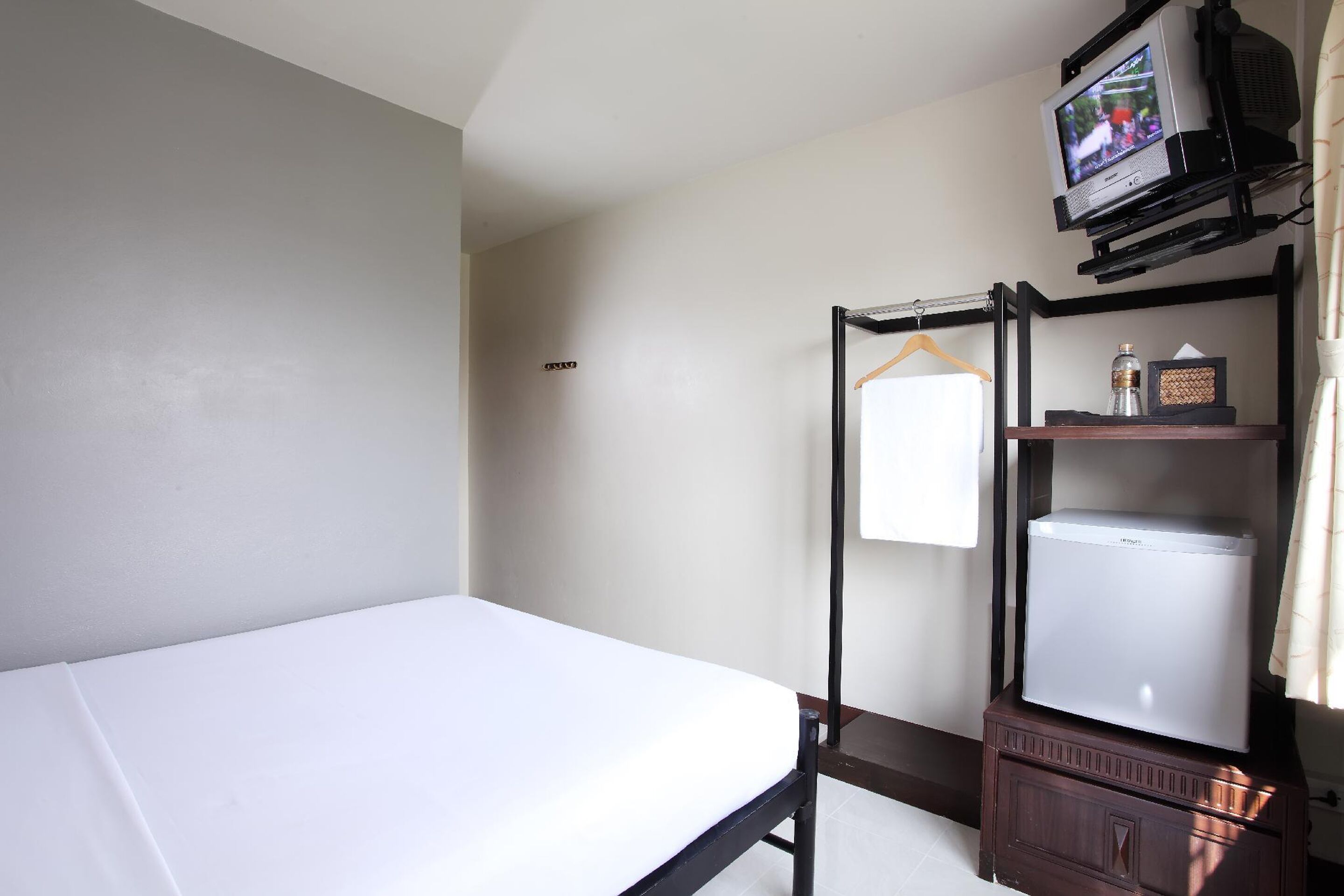 A single bed with a white mattress in a room with light-colored walls. There is a small wooden cabinet with a television mounted on a stand above it.