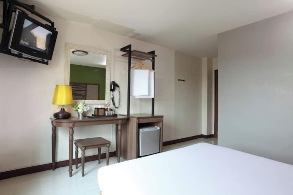 A hotel room with a bed, a desk with a lamp, a small refrigerator, a hanging coat rack, and a television mounted on the wall.