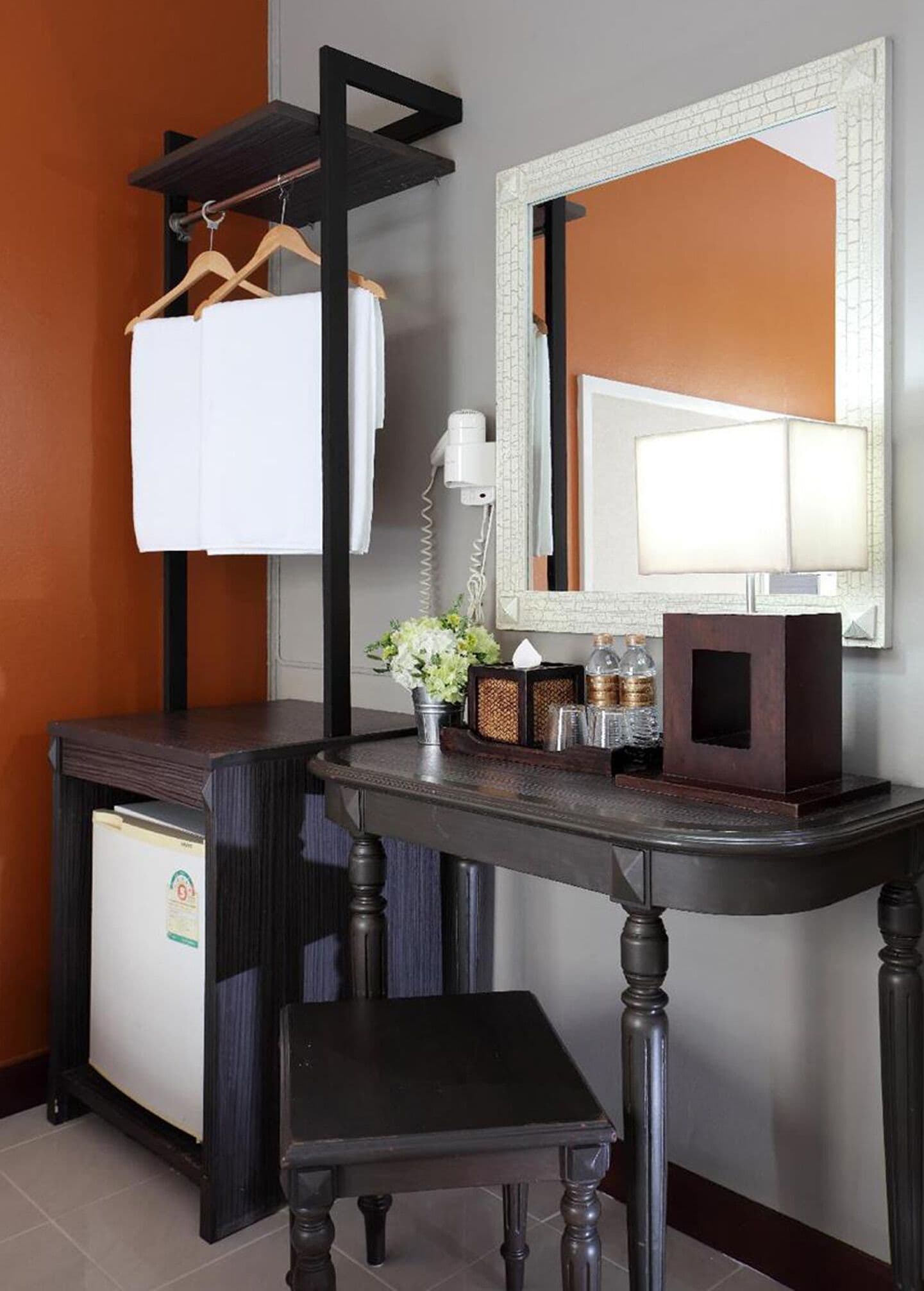 Habitación de hotel con un escritorio de madera oscura, una toalla blanca en un perchero, un secador de pelo, un espejo, una mesita con un jarrón de flores y un armario con refrigerador.