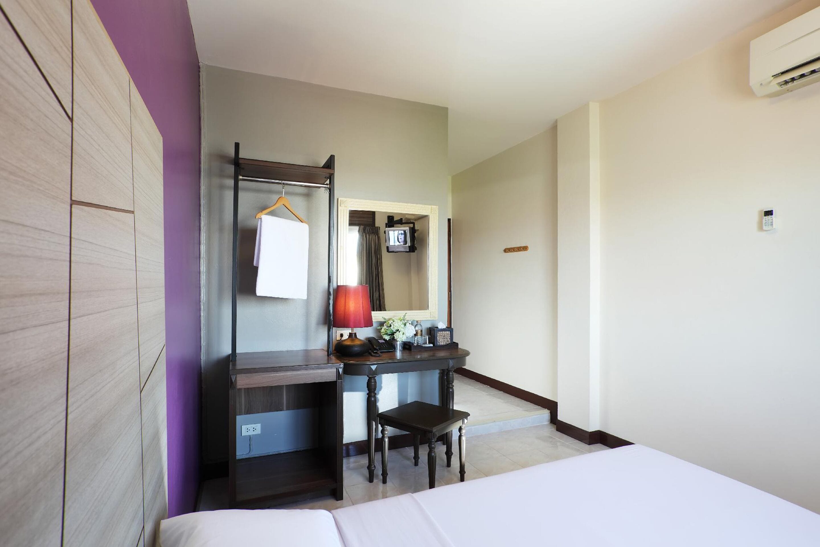 A hotel room with a bed, a desk with a lamp, a chair, a mirror, and an air conditioning unit.