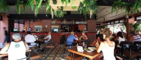 A restaurant with indoor plants hanging from the ceiling, tables and chairs arranged for dining, and a bar area in the background.