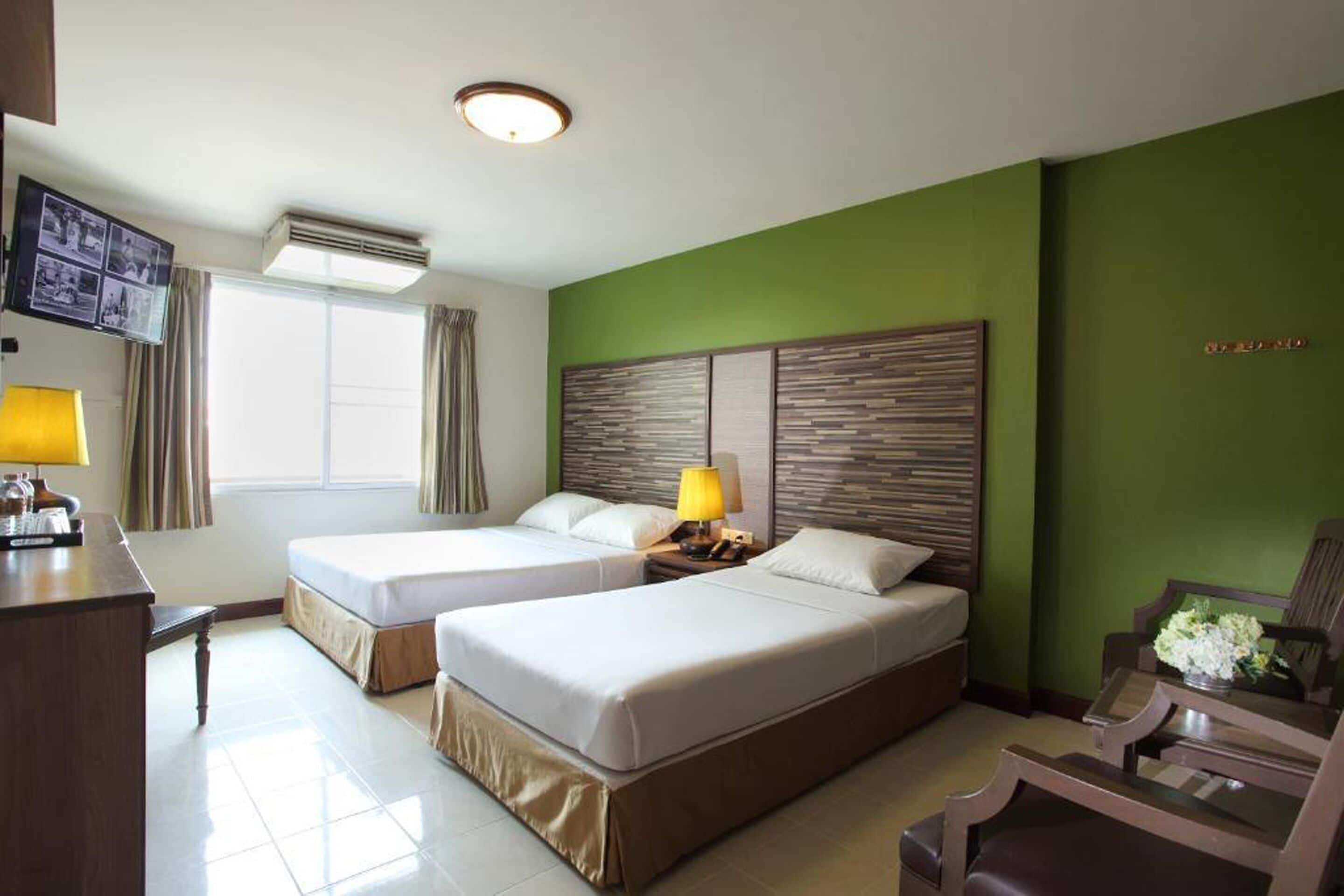 A hotel room with two beds, a desk with a lamp, a TV, and a green accent wall.