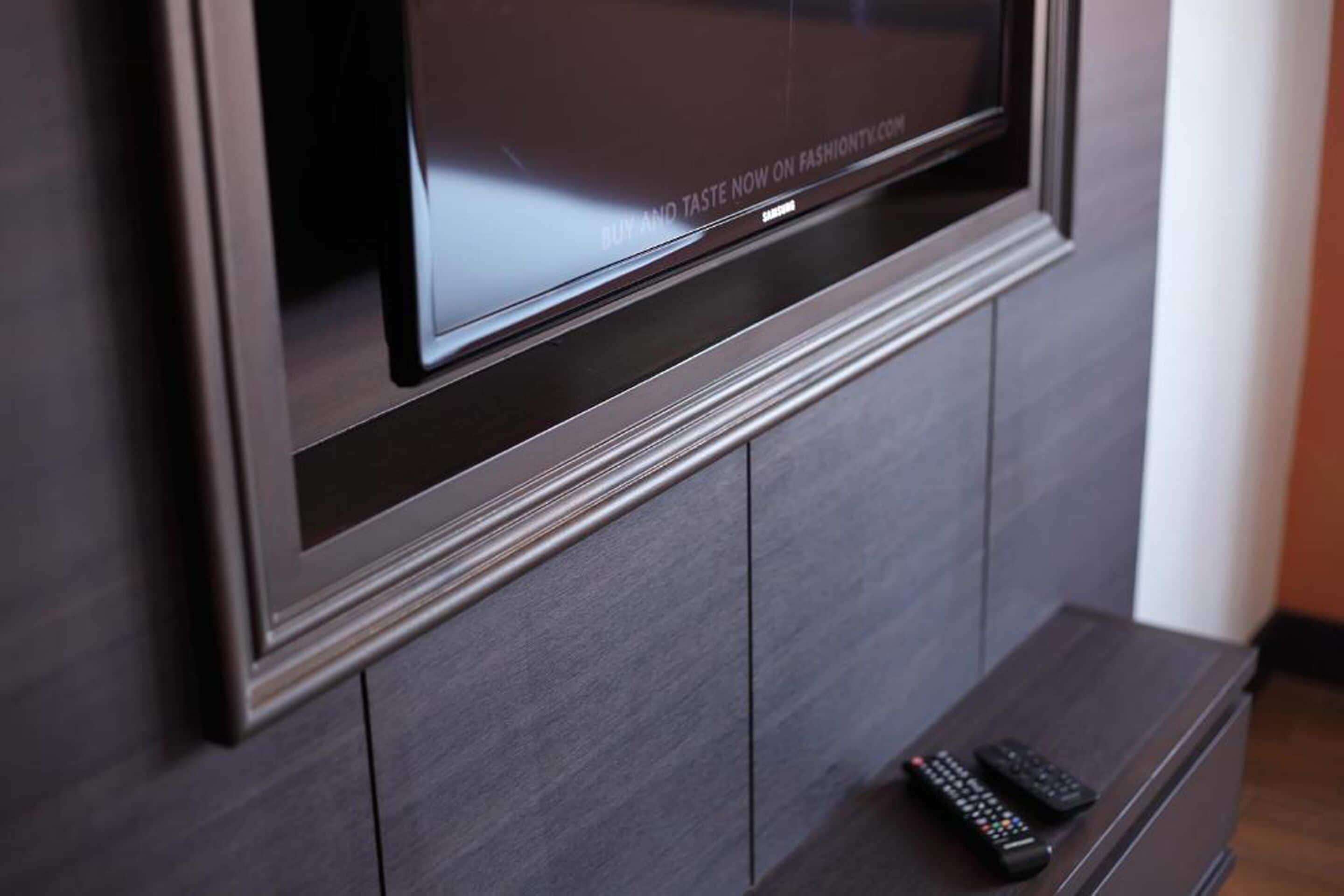 A flat-screen TV mounted on a dark wooden wall with a remote control on a small wooden stand below it.