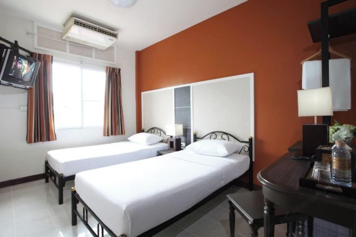 Habitación de hotel con dos camas individuales, un escritorio con lámpara, un televisor montado en la pared y una ventana con cortinas.