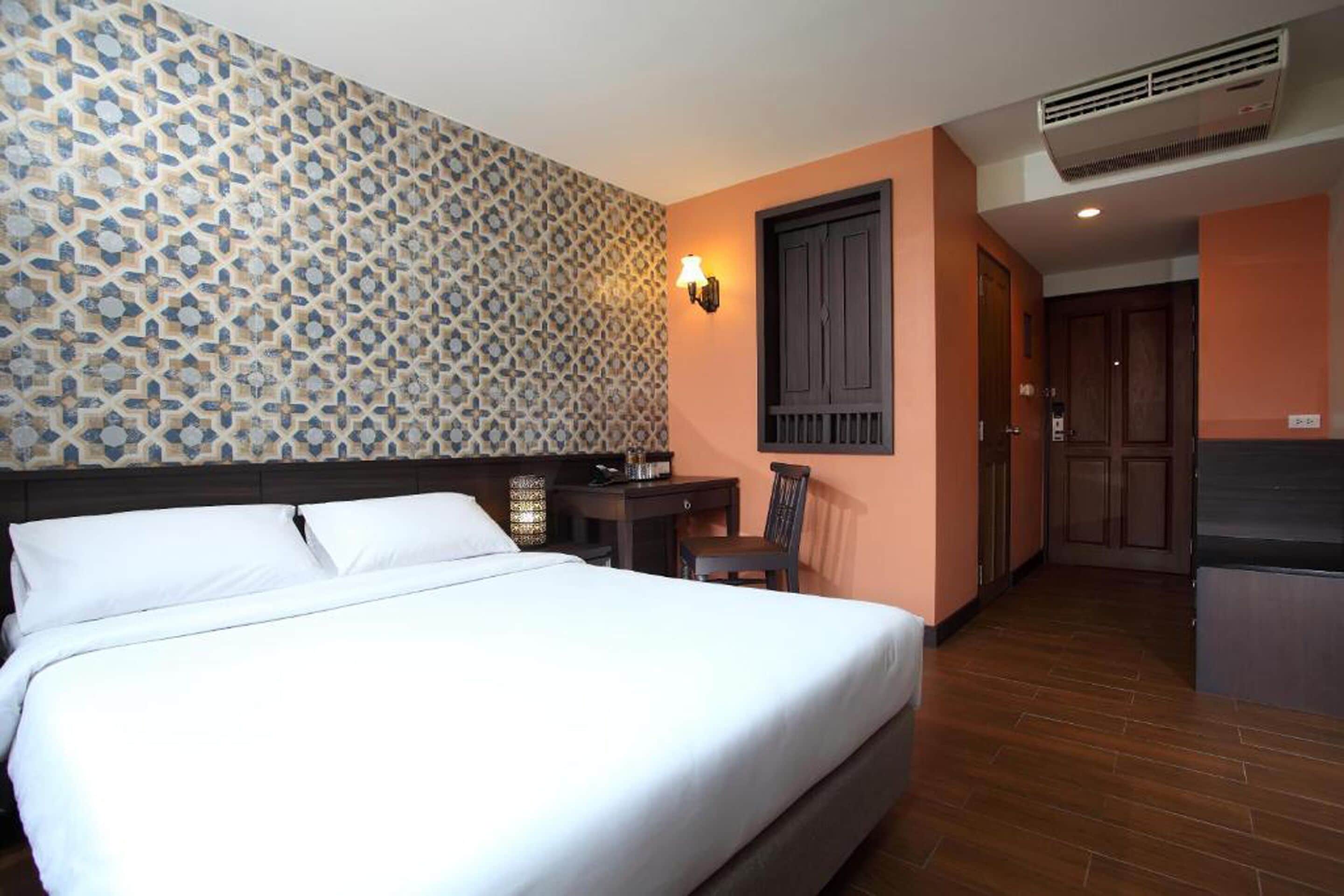 A hotel room with a bed, a desk, a chair, a wall-mounted lamp, and a door leading to a kitchenette.