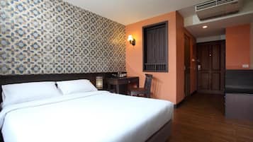 A hotel room with a bed, a desk, a chair, a wall-mounted lamp, and a door leading to a kitchenette.