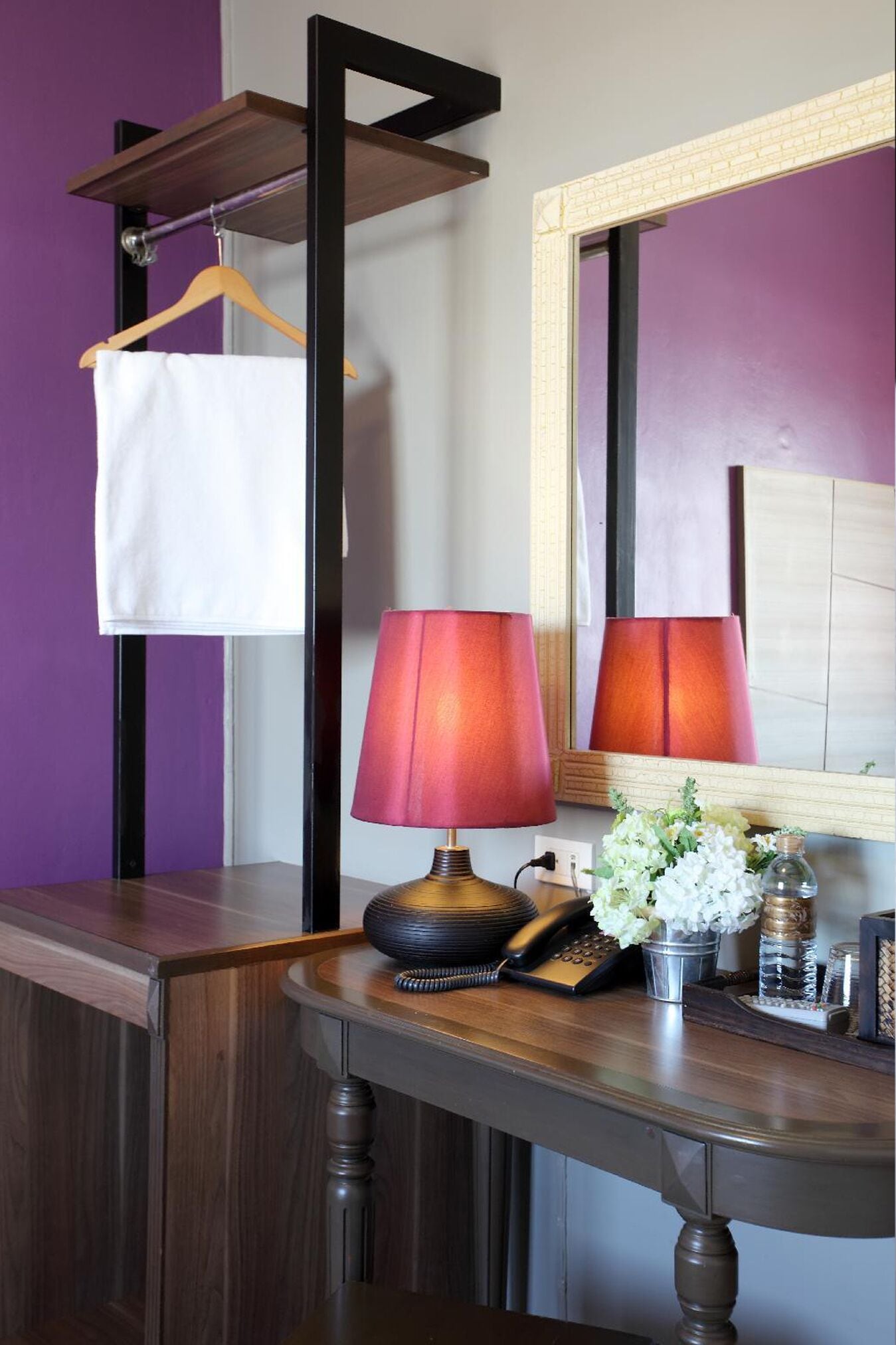A bathroom with a wooden vanity, a mirror, a red lamp, a white towel, and a small vase with white flowers.