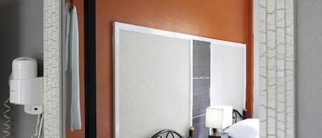 A hotel room with a large mirror, a bed with white linens, a bedside table with a lamp, and a hairdryer on the wall.
