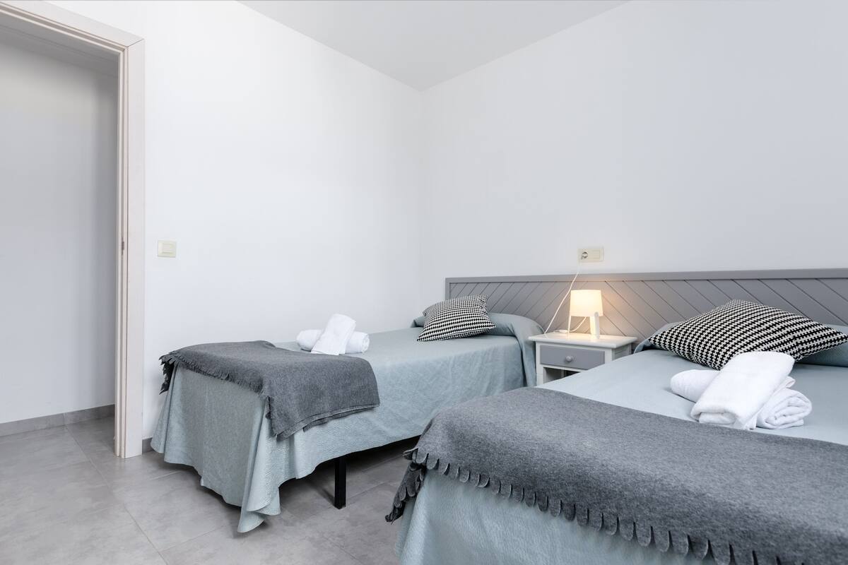 A room with two beds, each with a grey blanket and pillows, a bedside table with a lamp, and a door leading to another room.