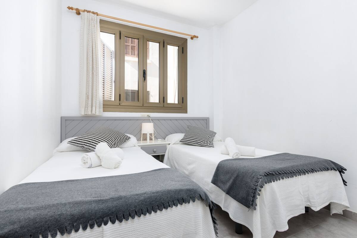 A hotel room with two beds, a window with curtains, a bedside table with a lamp, and a wall-mounted shelf.