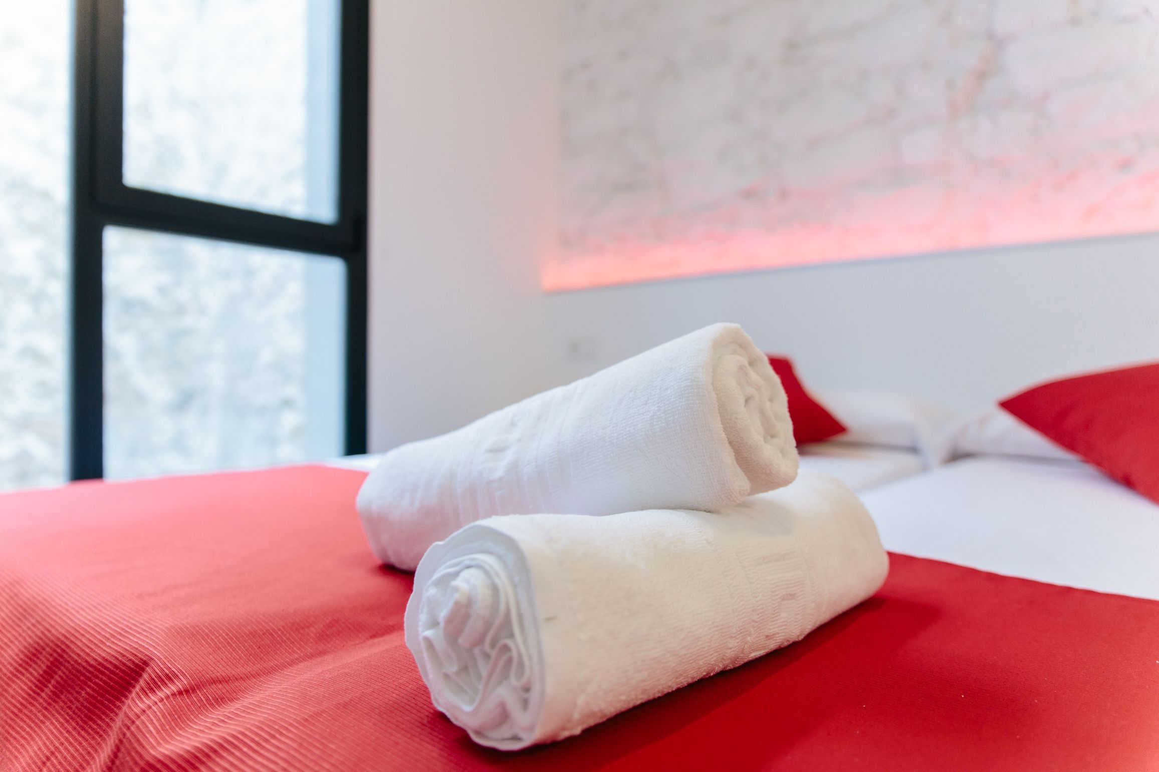 A neatly made bed with red bedding and two rolled white towels placed on it.