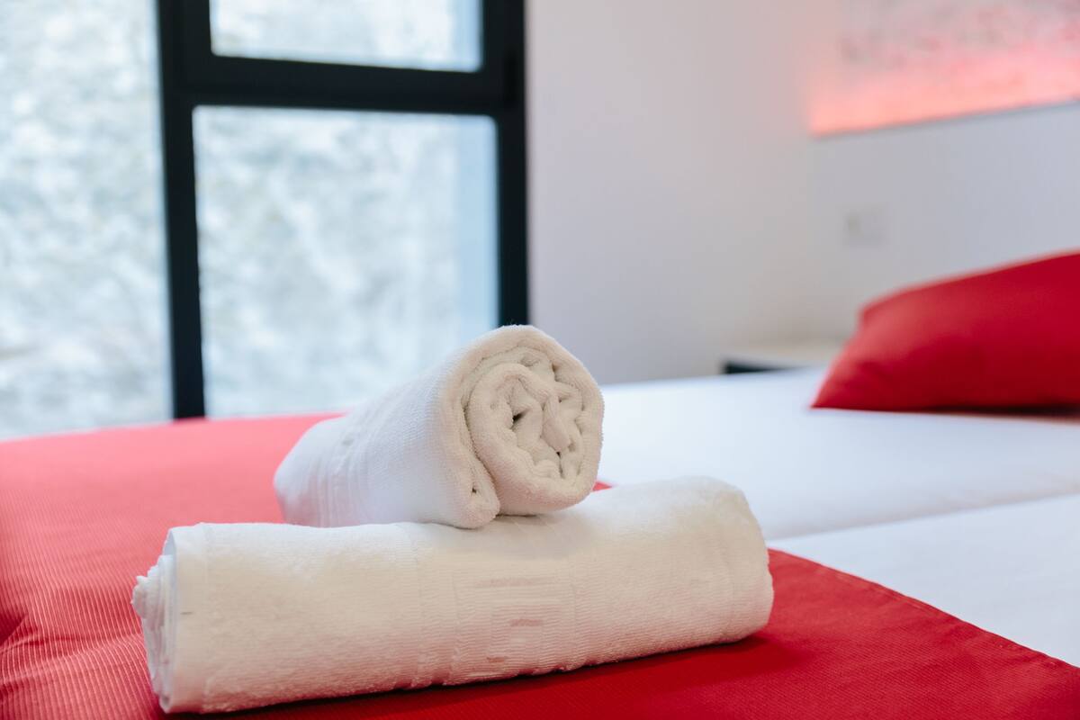 En prydligt vikt, vit handduk ligger på ett rött överkast i ett hotellrum, med ett fönster synligt i bakgrunden.