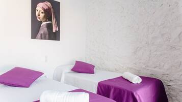 Una habitación de hotel con dos camas, cada una con una colcha morada y ropa blanca. Hay un cuadro en la pared sobre las camas.