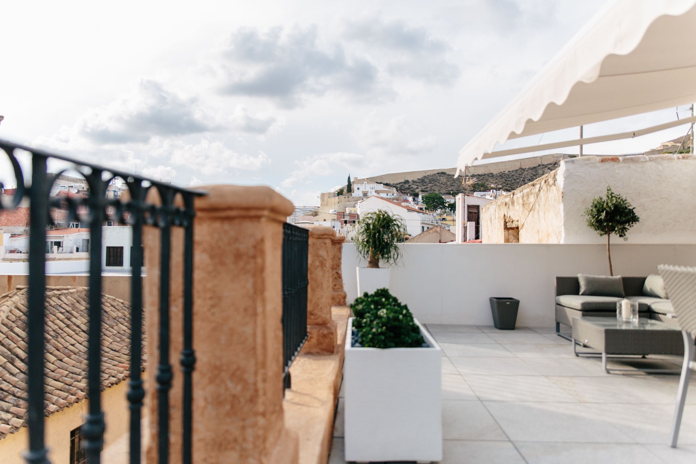 Una terraza en la azotea con barandilla blanca, una planta en maceta, mobiliario exterior y vista del paisaje urbano de fondo.
