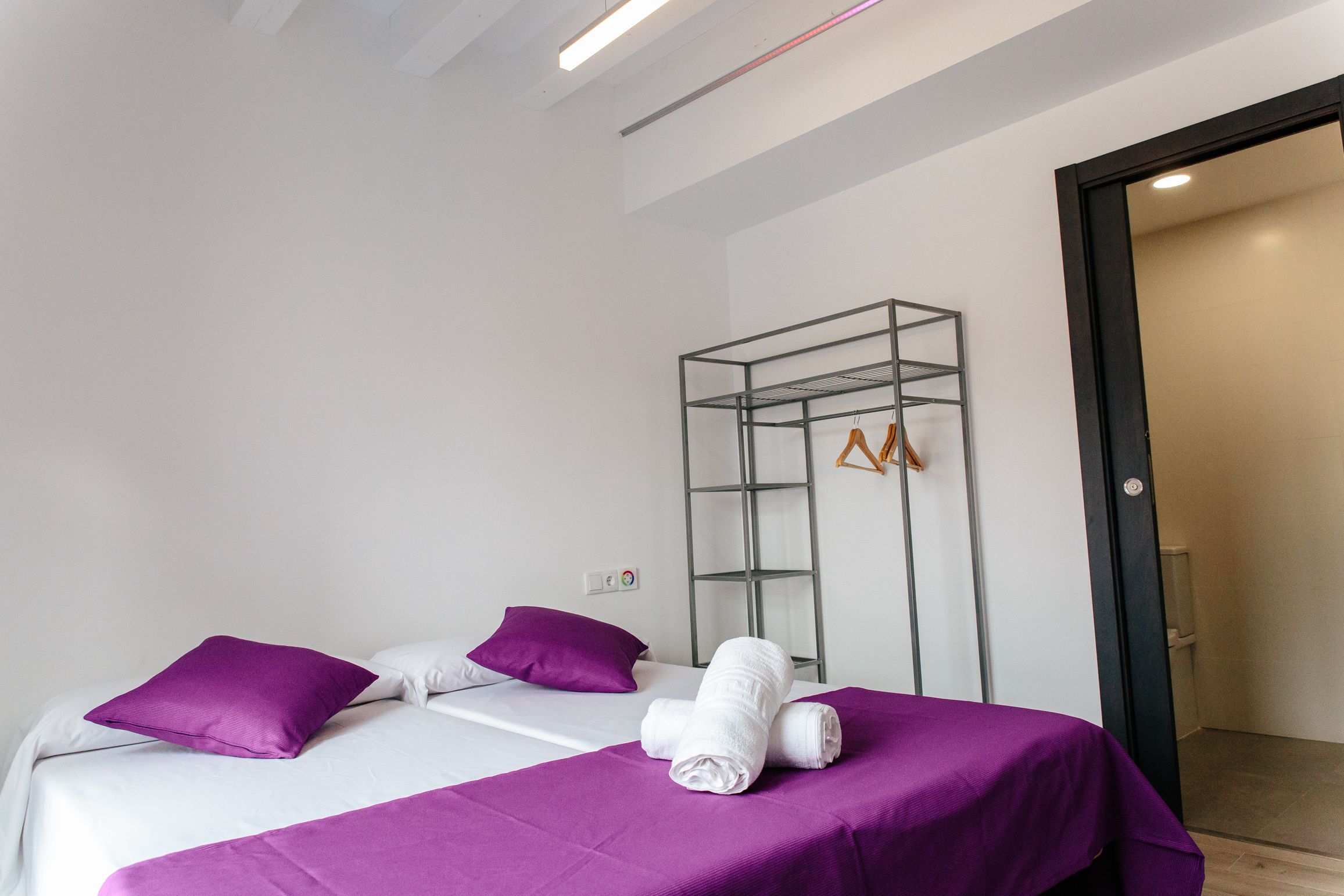 Quarto de hotel moderno com duas camas, roupa de cama roxa, uma estante de metal com roupas e uma porta que dá para o banheiro.