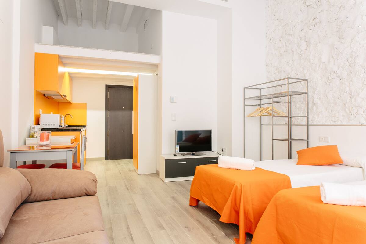 Ett modernt hotellrum med två sängar, en soffa, en TV och ett litet kök.