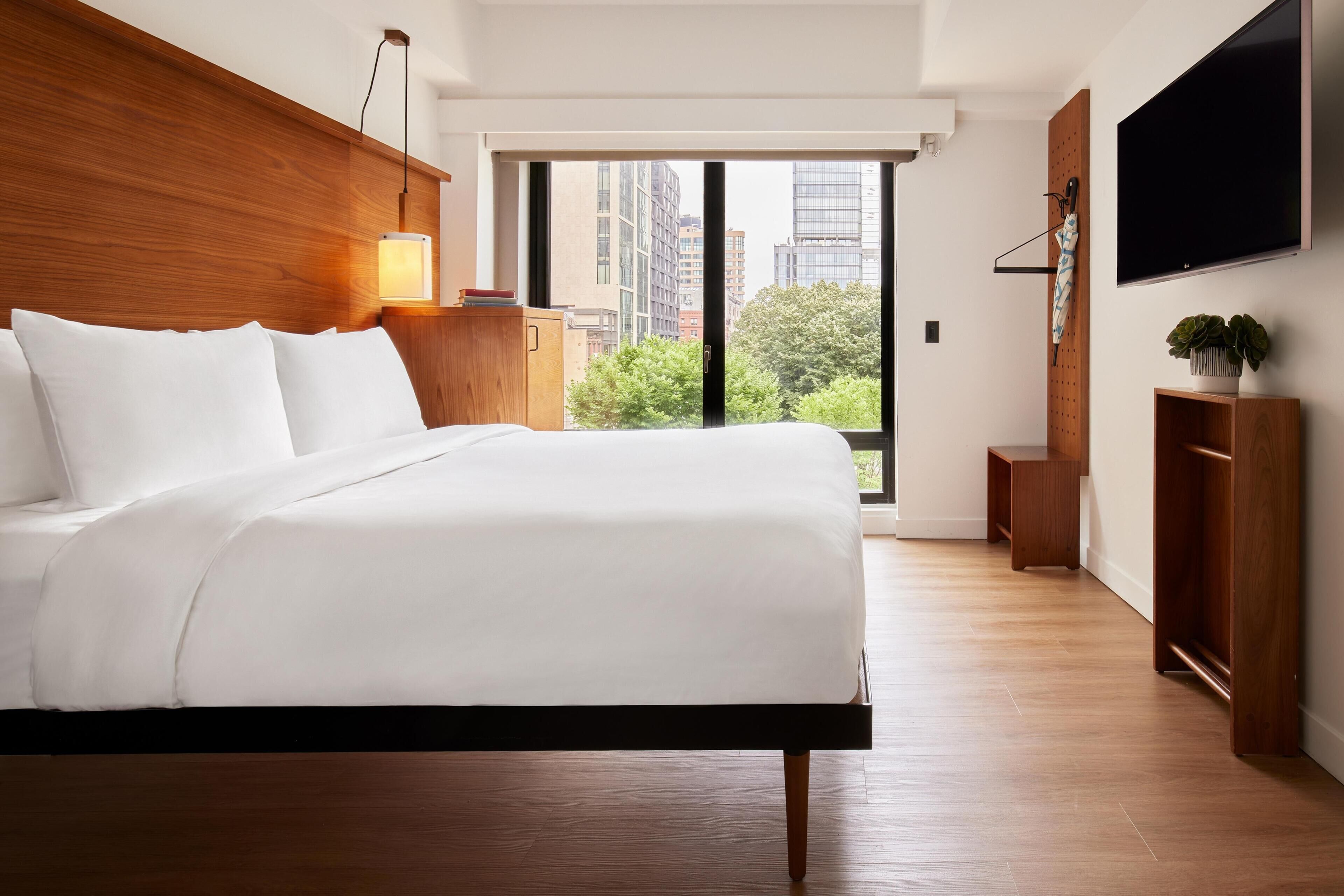 Habitación de hotel moderna con una cama grande, cabecera de madera, lámpara de noche, televisor de pantalla plana y vistas a la ciudad a través de un ventanal amplio.
