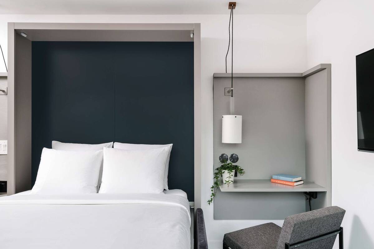 A modern hotel room with a large bed, a grey armchair, a small desk with a lamp, and a mounted TV.
