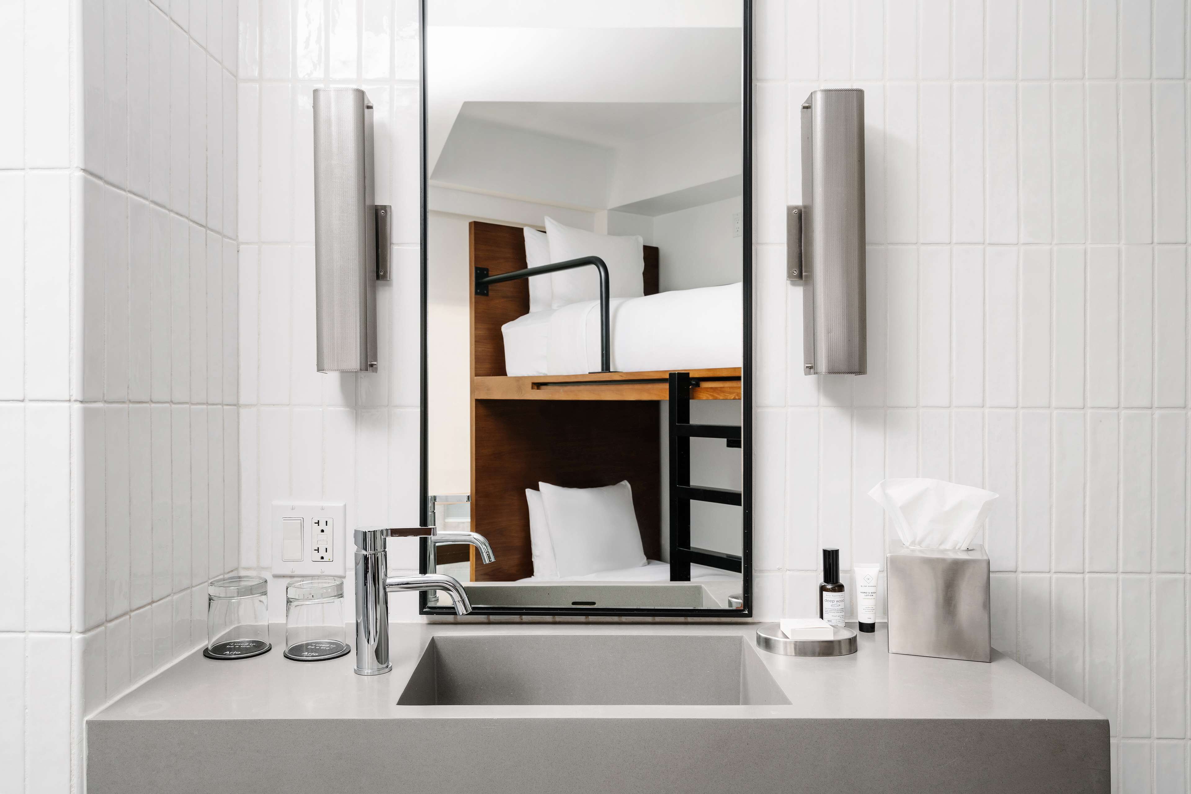 A modern bathroom with a large mirror, a sink, and a wooden bunk bed visible in the reflection.