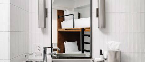 A modern bathroom with a large mirror, a sink, and a wooden bunk bed visible in the reflection.