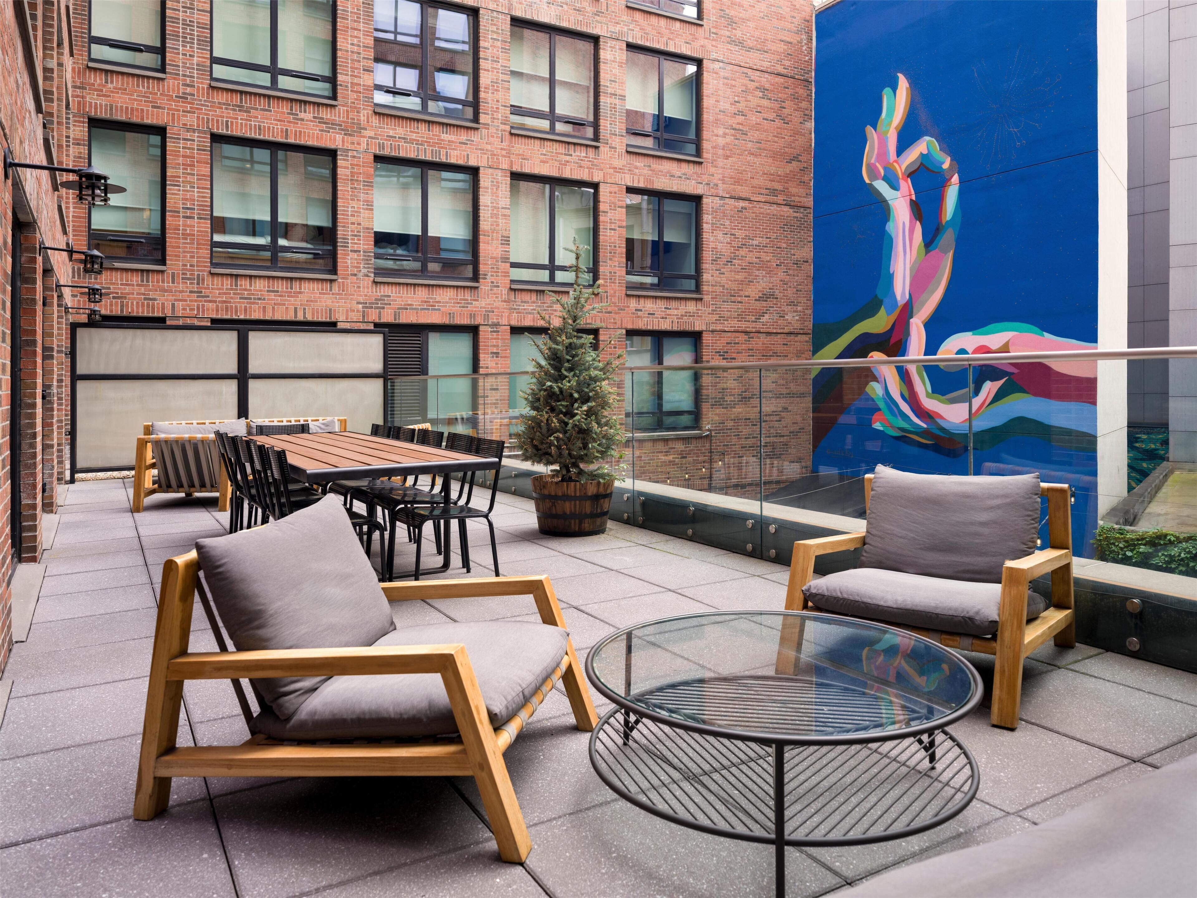An outdoor seating area with a table and chairs, a potted plant, and a mural on the building wall.