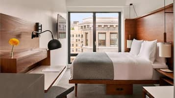 A modern hotel room with a large bed, a desk, a chair, and a view of the cityscape through a large window.