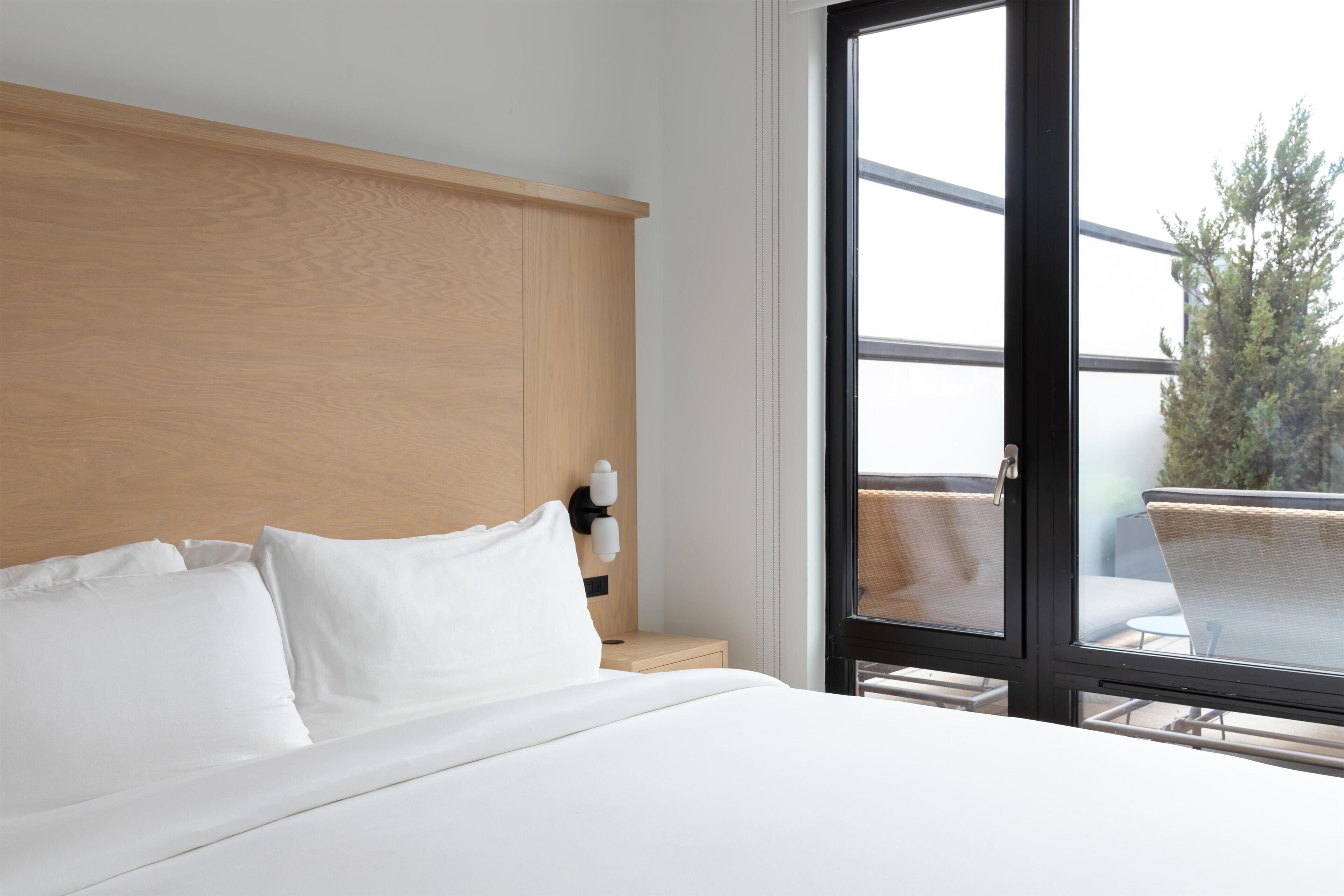 Habitación de hotel con una cama grande, cabecera de madera, mesita de noche con lámpara y vista al exterior a través de una puerta corredera.