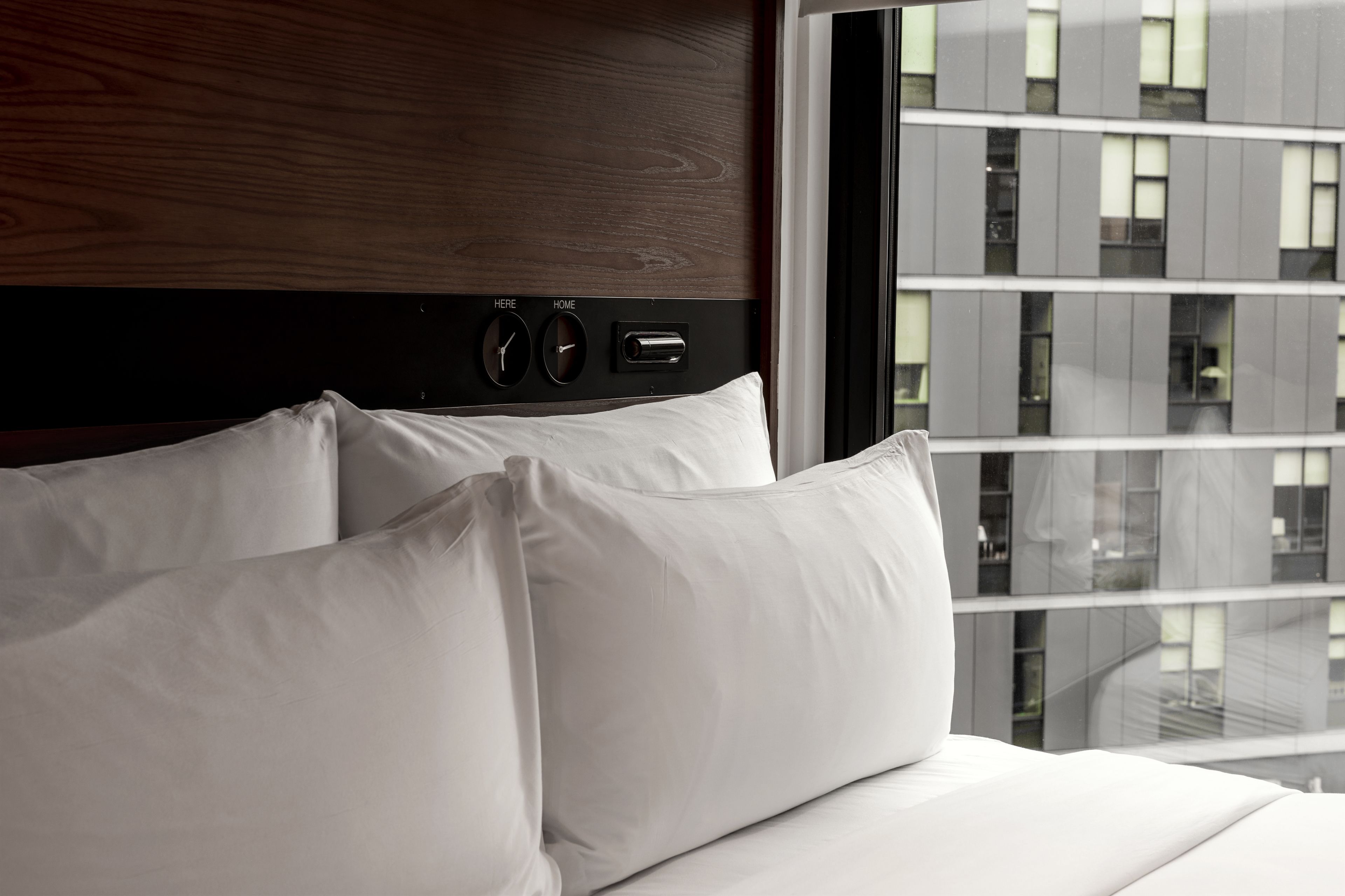 Une chambre d’hôtel avec une tête de lit en bois, un lit recouvert de draps blancs et une vue sur un immeuble moderne par la fenêtre.