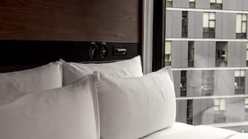 Une chambre d’hôtel avec une tête de lit en bois, un lit recouvert de draps blancs et une vue sur un immeuble moderne par la fenêtre.