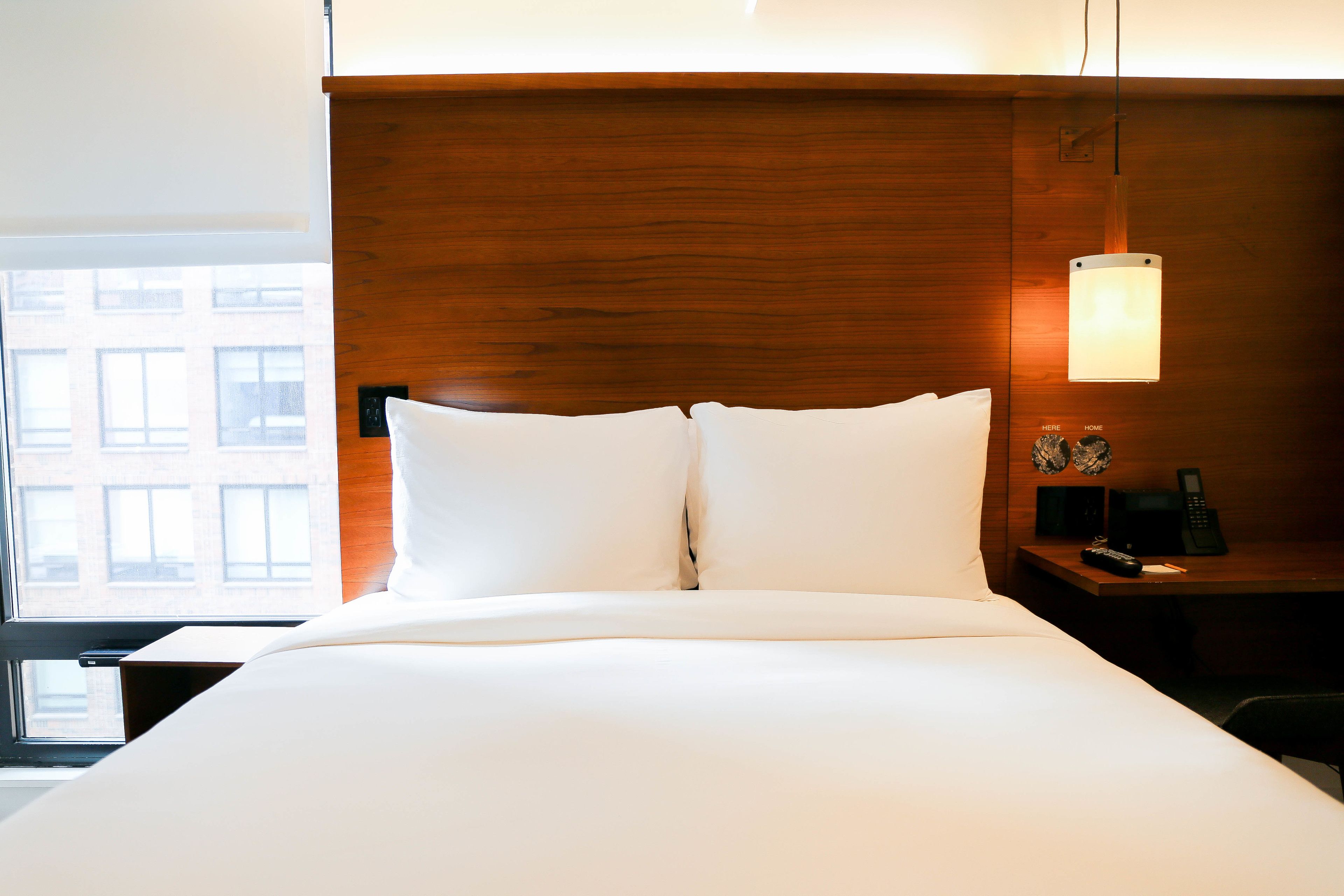 Ein Hotelzimmer mit einem großen Bett, einem Holzkopfteil, einem Nachttisch mit Lampe und Blick auf Gebäude durch das Fenster.