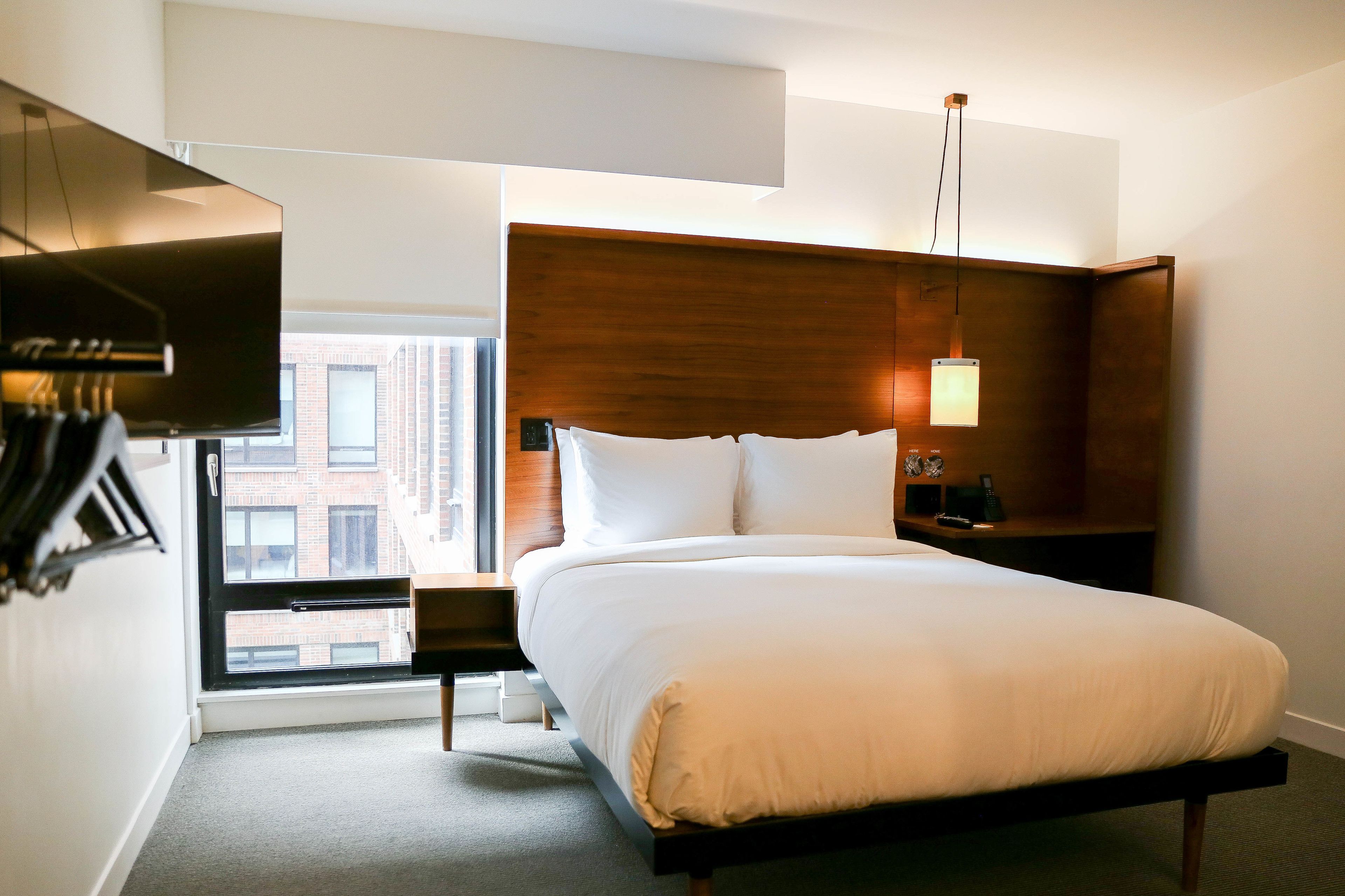 Una habitación de hotel moderna con una cama grande, un cabecero de madera, una mesita de noche, una lámpara y vistas a un edificio a través de una ventana.